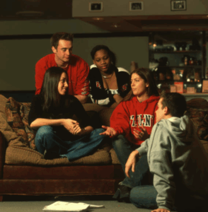 Four university students in casual clothes sit and talk together on sofas in a lounge area. Papers and notebooks are spread on the floor in front of them, suggesting a relaxed group study or discussion session.